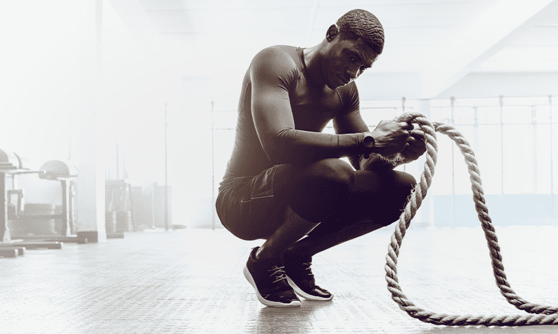 A man in athletic wear is crouching in a gym, holding a thick rope with both hands