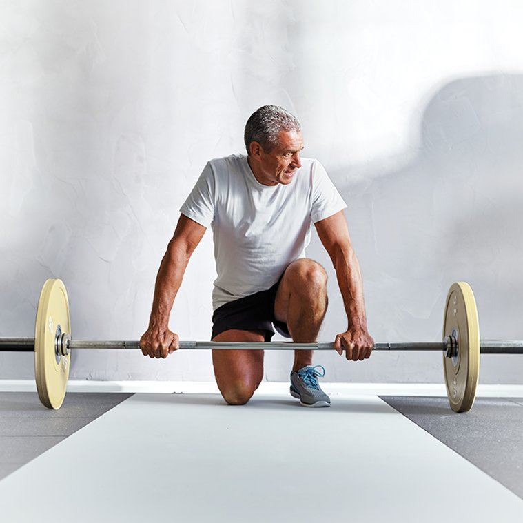 An older man is kneeling on one knee while holding a barbell with weights