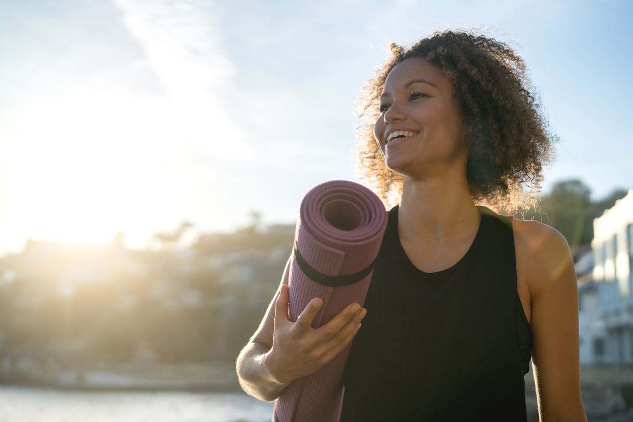 A woman smiling, holding a rolled-up yoga mat at sunset